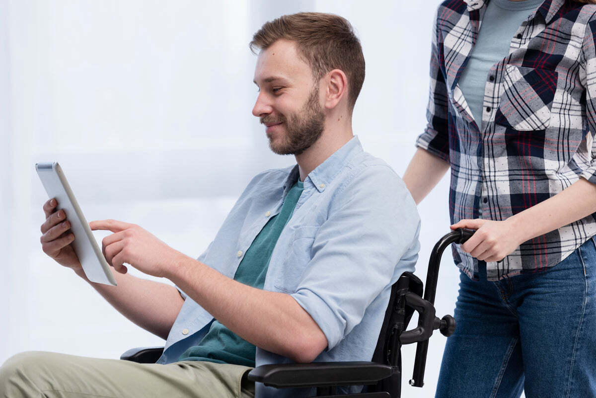 A man in a wheelchair using a tablet as someone stands behind him